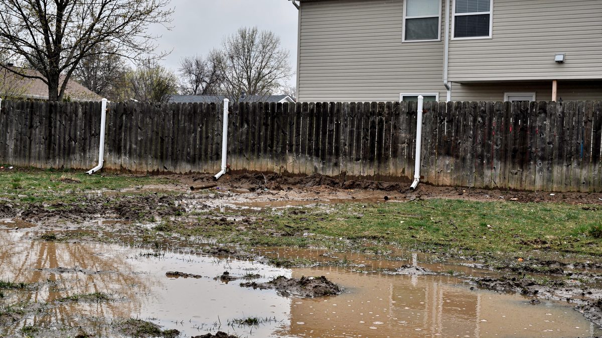 Standing water pooled across a saturated back lawn a day after heavy rain at a Kansas City metro home, downspouts discharging straight onto the soil at the foundation